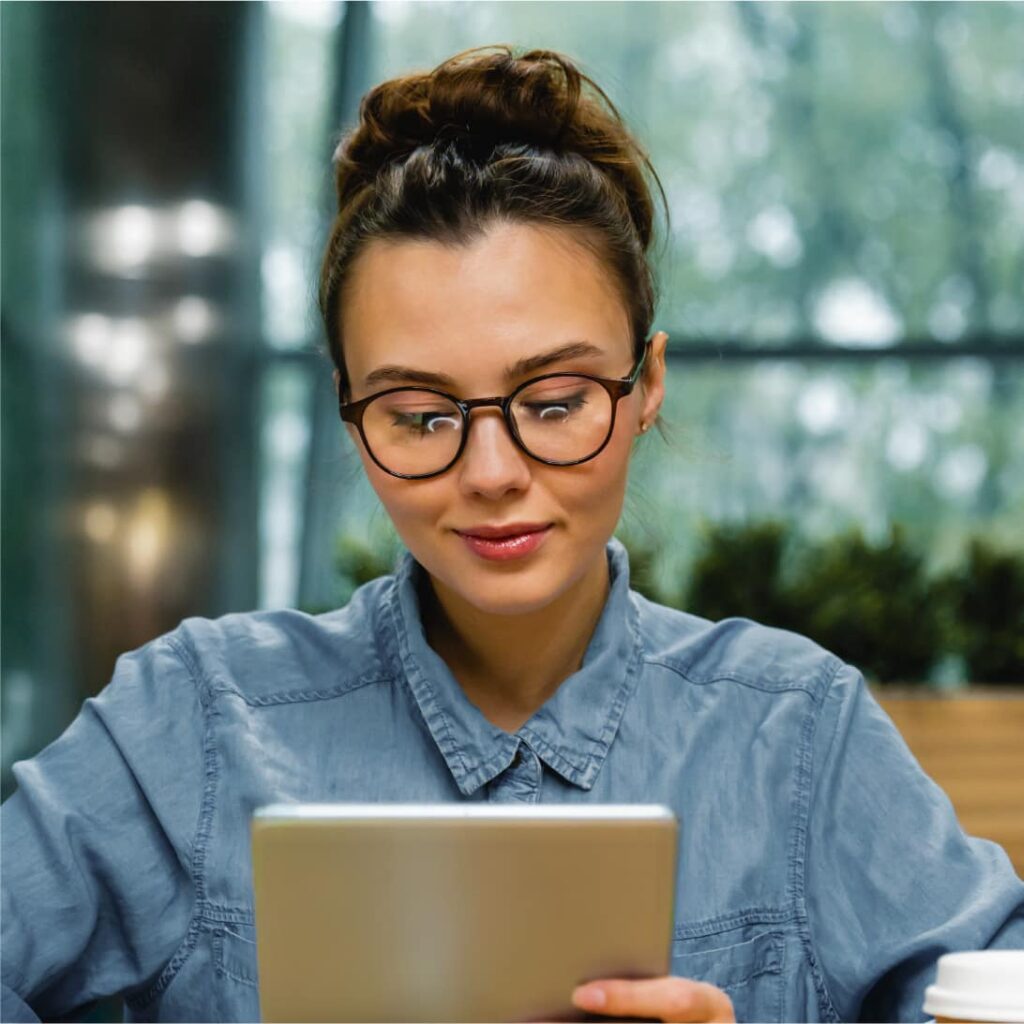 Una persona con gafas y un peinado de moño está mirando la pantalla de una tableta, sentada frente a un fondo interior borroso.