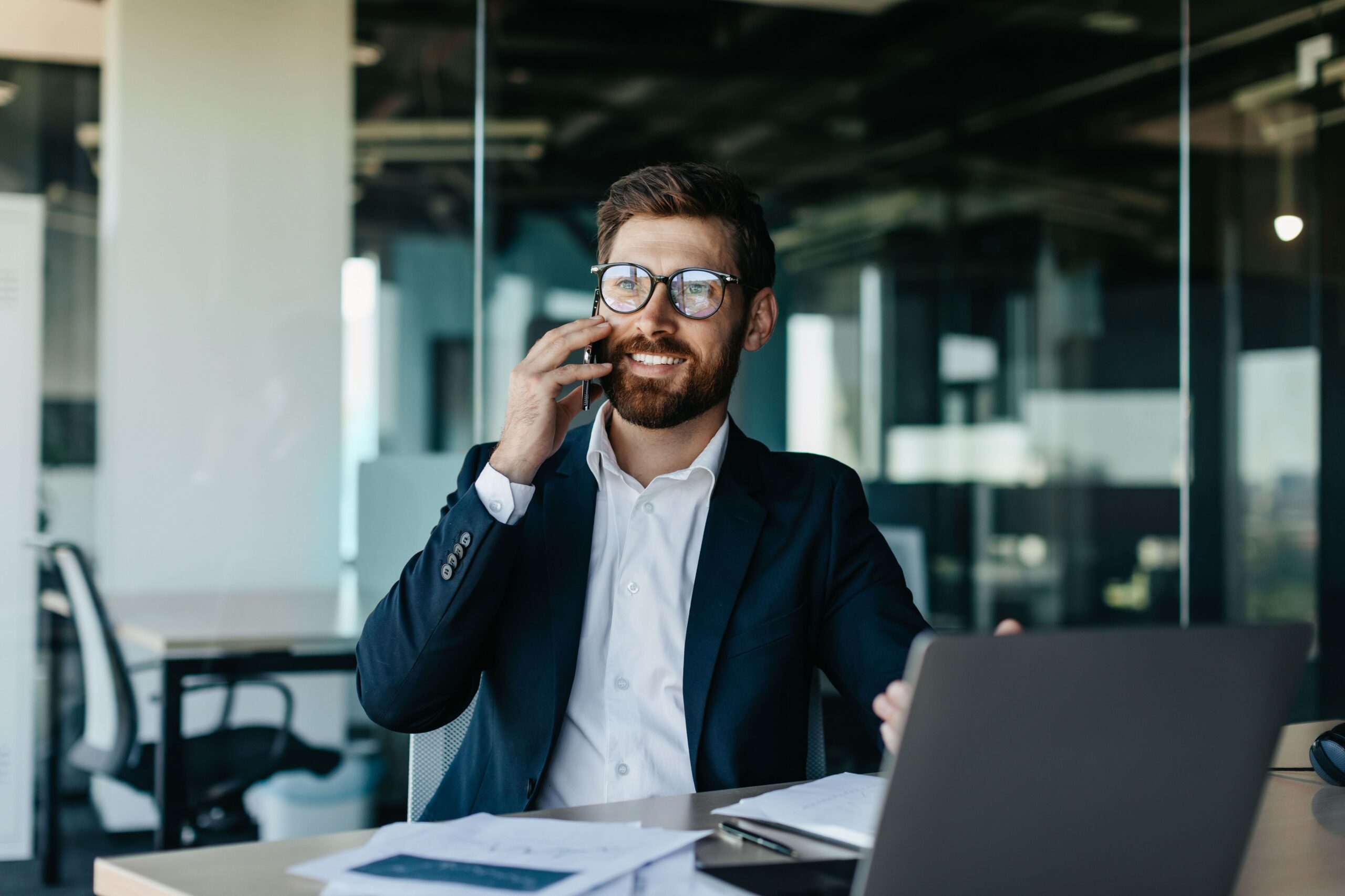 Un hombre con traje y gafas habla por teléfono mientras está sentado en su escritorio con una computadora portátil y documentos en un ambiente de oficina.
